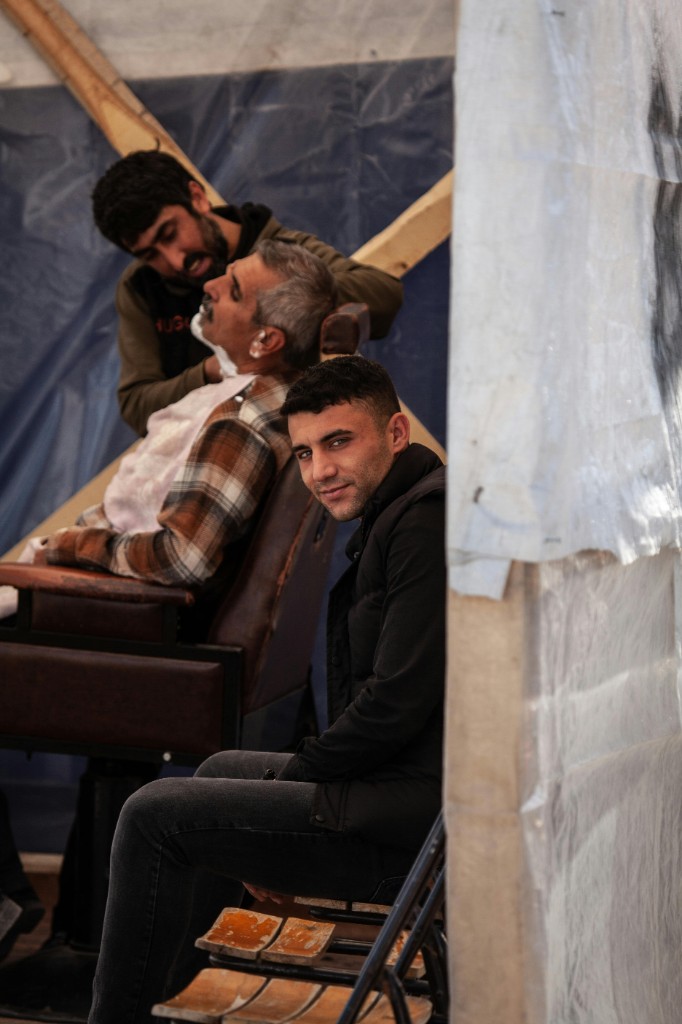 Street barbershop: a barber shaving a reclined customer in a leather chair under a blue tarp; another man sits in the foreground.
