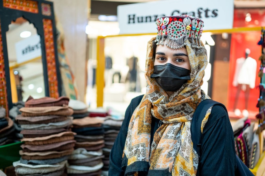 A woman in traditional dress and jewelry stands in a bright boutique with stacked wool hats and crafts on display.