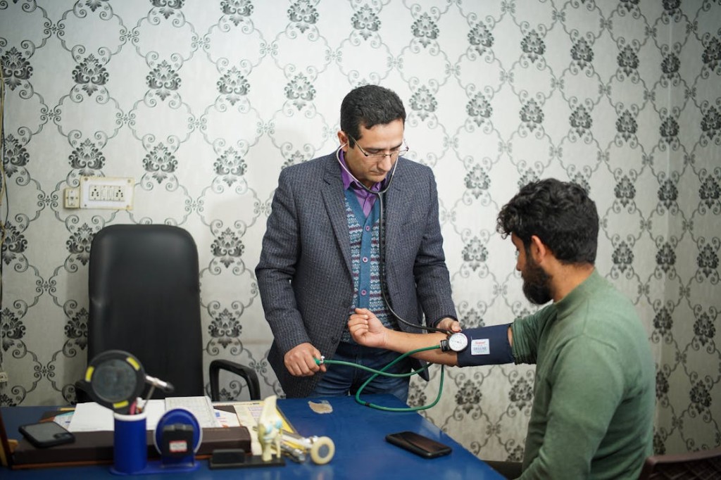 A healthcare provider measures a seated patient’s blood pressure with a stethoscope and manual cuff at a clinic desk.