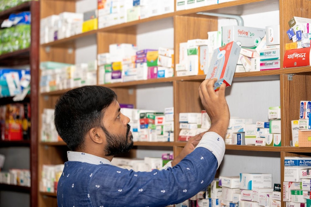 A pharmacist reaches for a medicine box from densely stocked wooden shelves in a bright pharmacy.