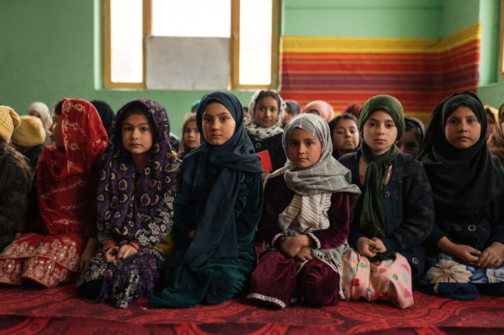 Young students in headscarves sit in rows on a patterned carpet in a classroom, facing the camera in soft natural light.